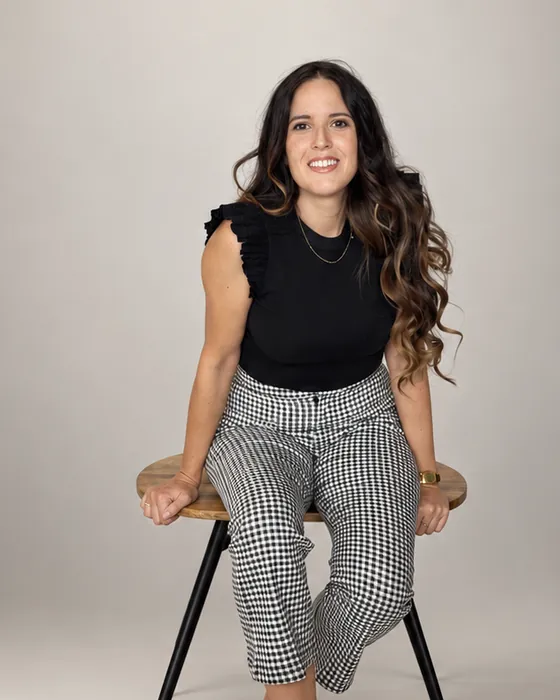 Mónica Pérez sentada en un taburete, inclinada hacia delante en actitud de escucha, en un estudio de fondo claro.