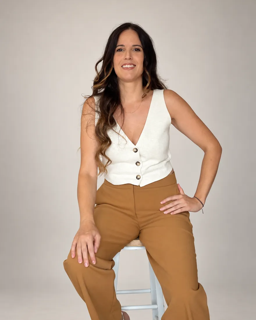 Mónica Pérez sonriendo, sentada en un taburete, con chaleco blanco y pantalón camel, sobre un fondo claro de estudio.